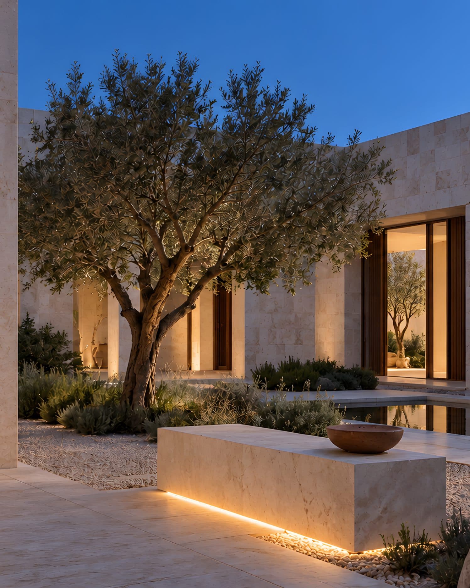 The same limestone courtyard at dusk: lamps under the colonnade and the bench, the olive tree against a deep blue sky